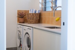 A laundry area with two front-loading washing machines, a wicker basket with towels, and a window above the sink. - The Lighthouse (Airlie Beach)