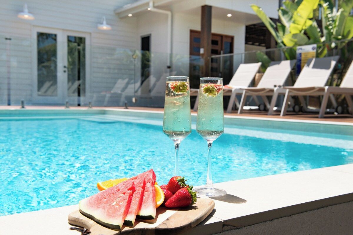 A poolside setting with two glasses of a clear beverage, a wooden board with sliced watermelon and strawberries, and a pool with lounge chairs in the background.