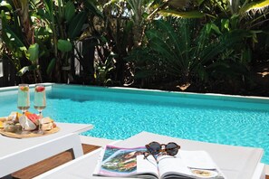 Escena junto a la piscina con una mesa, una bandeja de comida y una revista abierta.