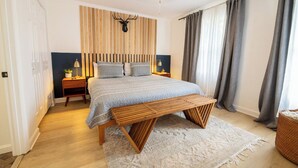 Ein Schlafzimmer mit einem Bett, einer hölzernen Bank, einem Kopfteil mit vertikalen Lamellen, einem an der Wand befestigten Hirschkopf und einem Nachttisch mit Lampe.