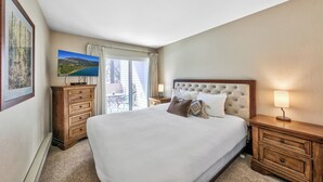 A hotel room with a large bed, wooden nightstands, a flat-screen TV, a window with curtains, and a view of a landscape.