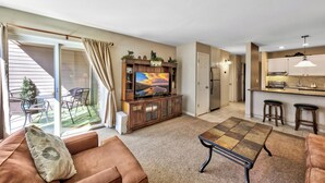A living room with a sofa, a coffee table, and a TV on a wooden cabinet. There is a sliding glass door leading to a patio with outdoor furniture.