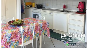Una cucina con mobili bianchi, un fornello, un lavello e una macchina per caffè. Si nota un tavolo con una tovaglia colorata e una ciotola di frutta.