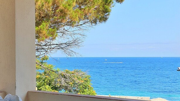 Un balcone con una sedia bianca, un tavolino con una pianta e un bicchiere, e vista sul mare.