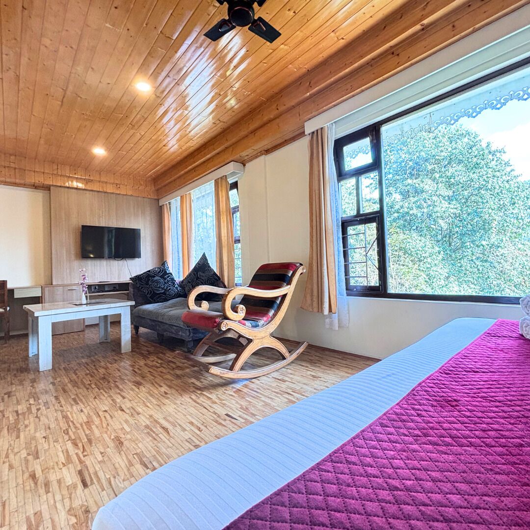 Et værelse med trælofter, en seng med et pink quilt, en gyngestol, en sofa med mønstrede puder, et fjernsyn på væggen og store vinduer med udsigt til træer.