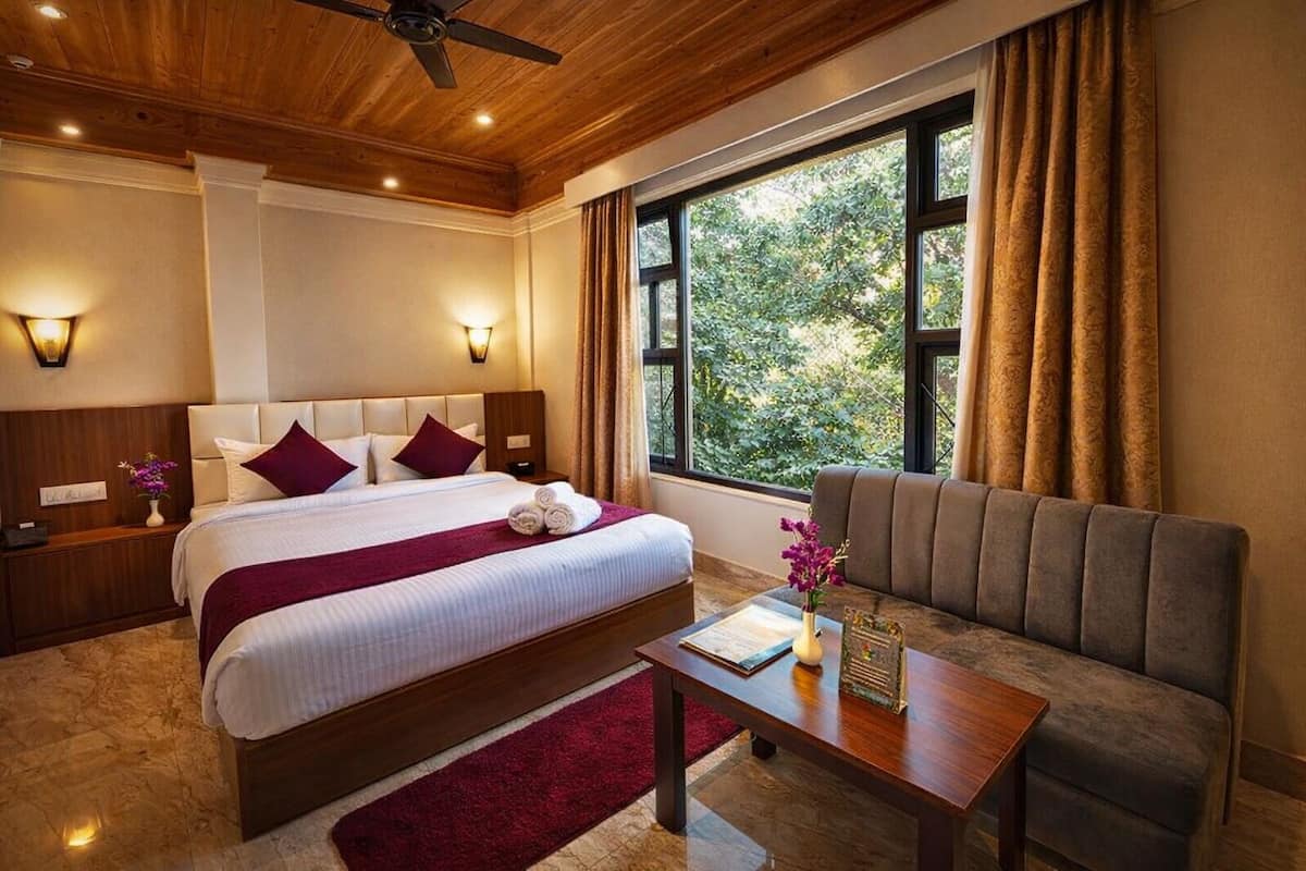 A hotel room with a bed, a sofa, a window with curtains, a ceiling fan, and a nightstand with a vase.