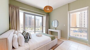 A bedroom with a large bed, a bedside table, a mirror, and a view of tall buildings through large windows. - The Point Tower - Vibrant One Bed in Marina (Dubai Marina)