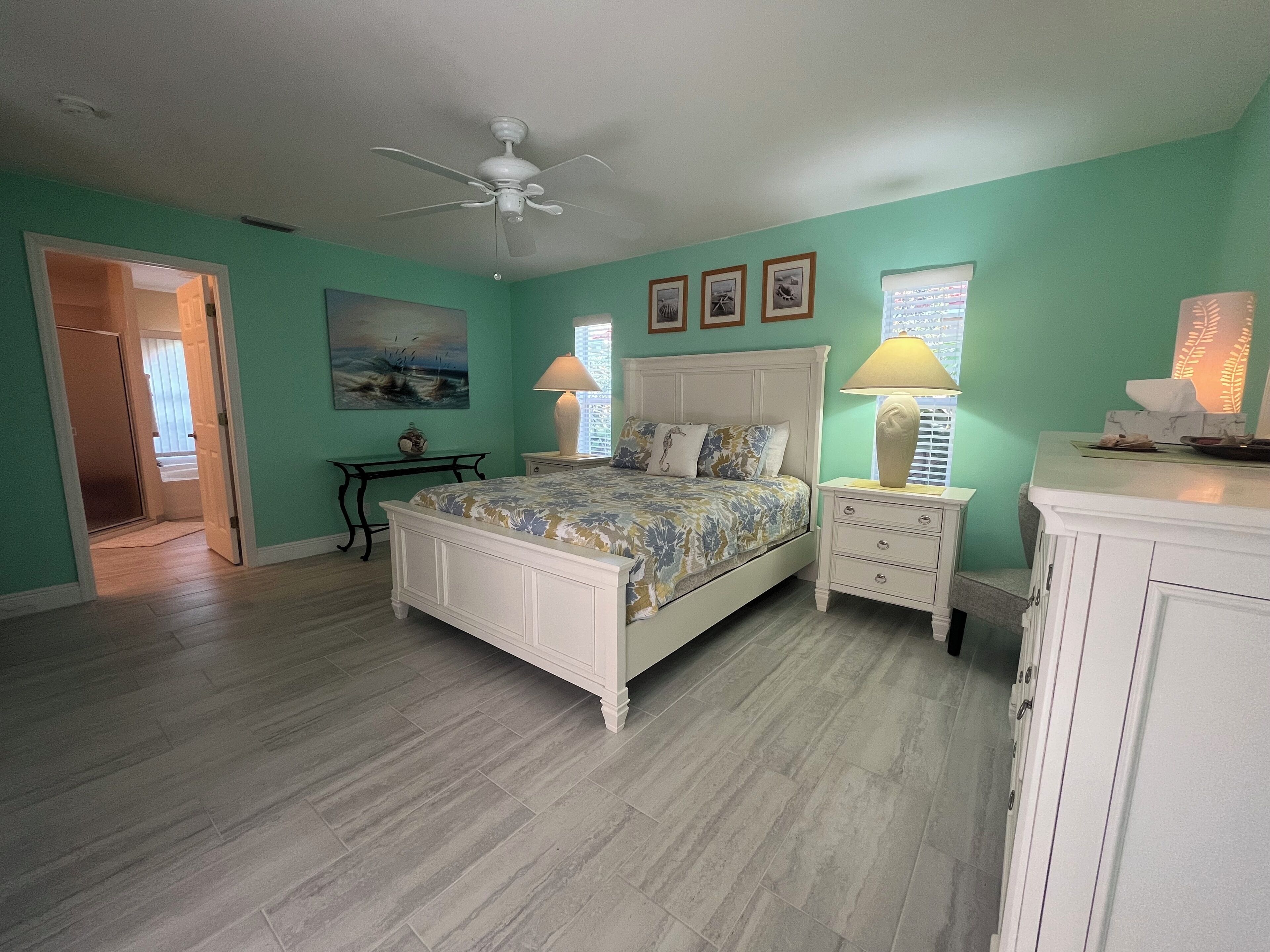Une chambre à coucher comprenant un lit, des tables de chevet, un ventilateur de plafond, un tableau et une fenêtre avec des stores.