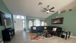 A spacious living room with a dark green wall, a ceiling fan, a black entertainment unit, and a large window with a view of greenery. - The White Egret, Private heated Pool, Fresh Water Canal, Beach, Fishing, WIFI (Cape Coral)