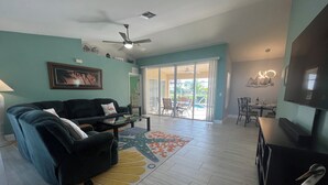 A living room with a sofa, coffee table, and a flat-screen TV. There is a ceiling fan, a dining area with chairs, and a large window with a view of the outdoors. - The White Egret, Private heated Pool, Fresh Water Canal, Beach, Fishing, WIFI (Cape Coral)