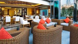 Un área de estar moderna en interiores con mobiliario de mimbre y cojines naranja, con un muro de cristal que ofrece vistas a la ciudad.