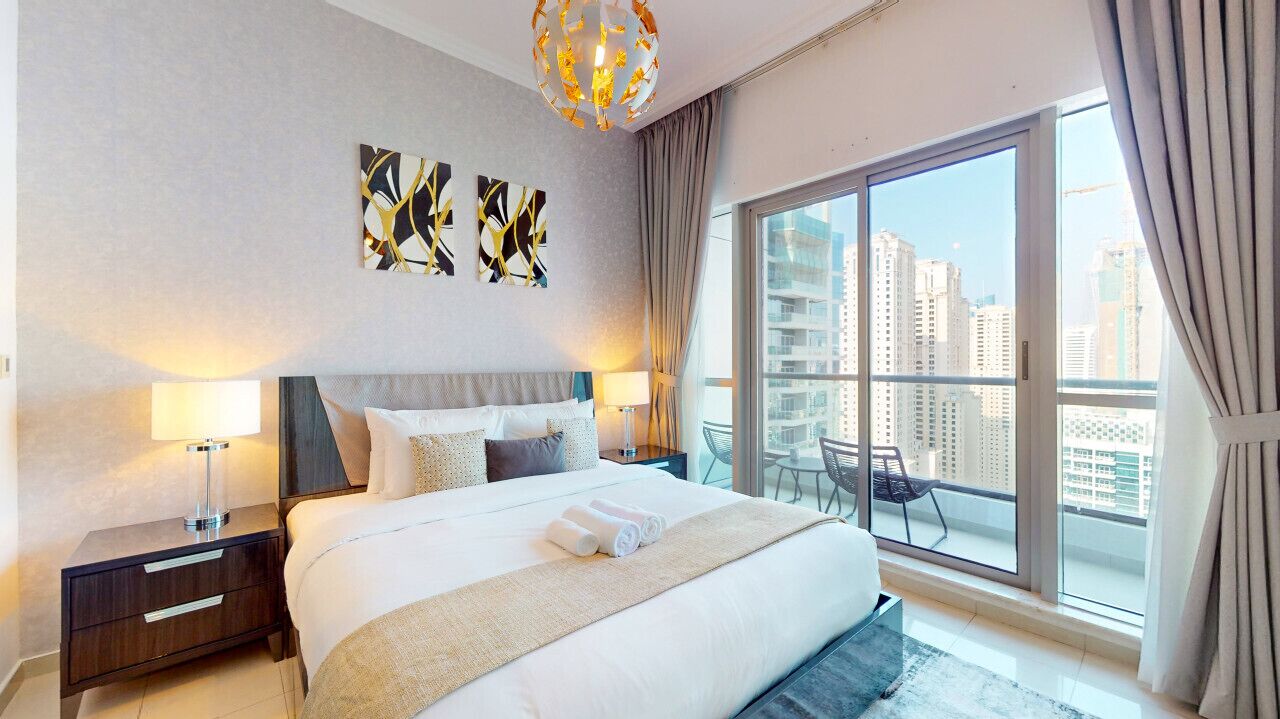 Quarto de hotel moderno com uma cama grande, duas mesas de cabeceira, um criado-mudo, uma varanda com vista para a cidade e arte abstrata na parede.