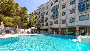 Una piscina con sillas de descanso y sombrillas, rodeada por un edificio con balcones y ventanas.