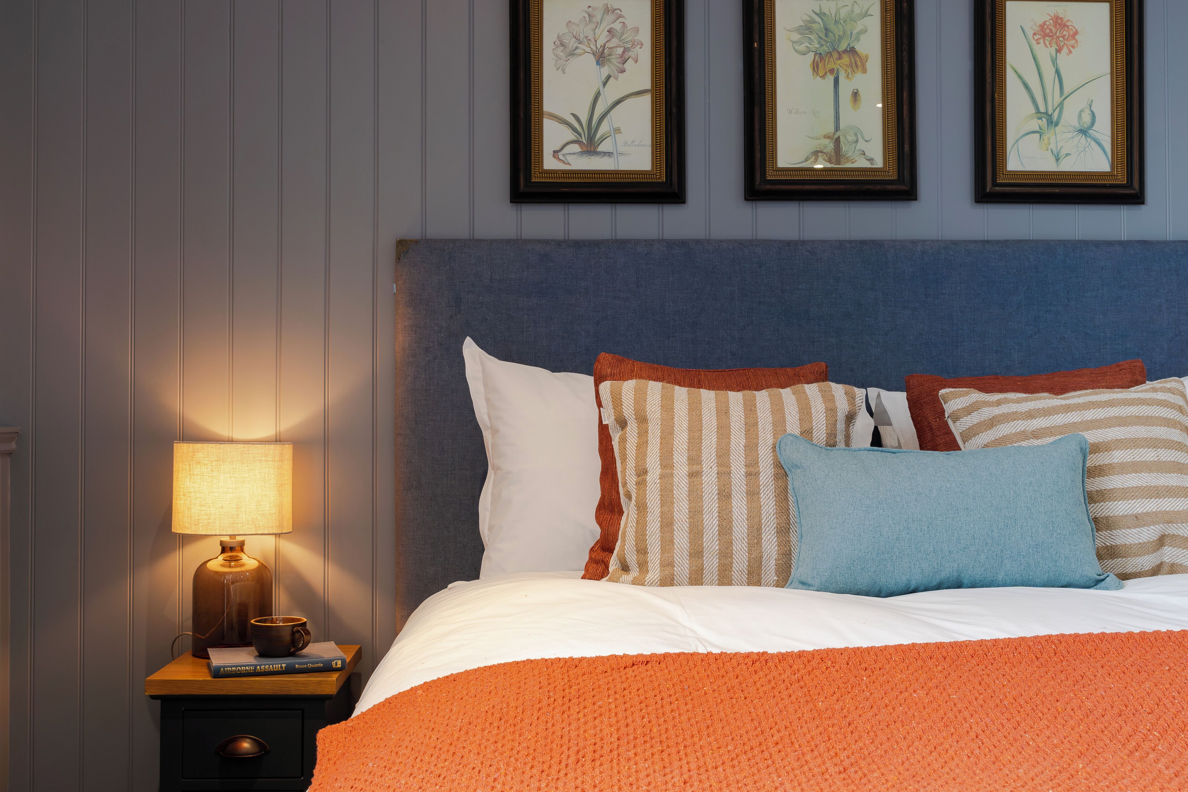A neatly made bed with a blue headboard, a wooden nightstand with a lamp, and framed botanical artwork on the wall.