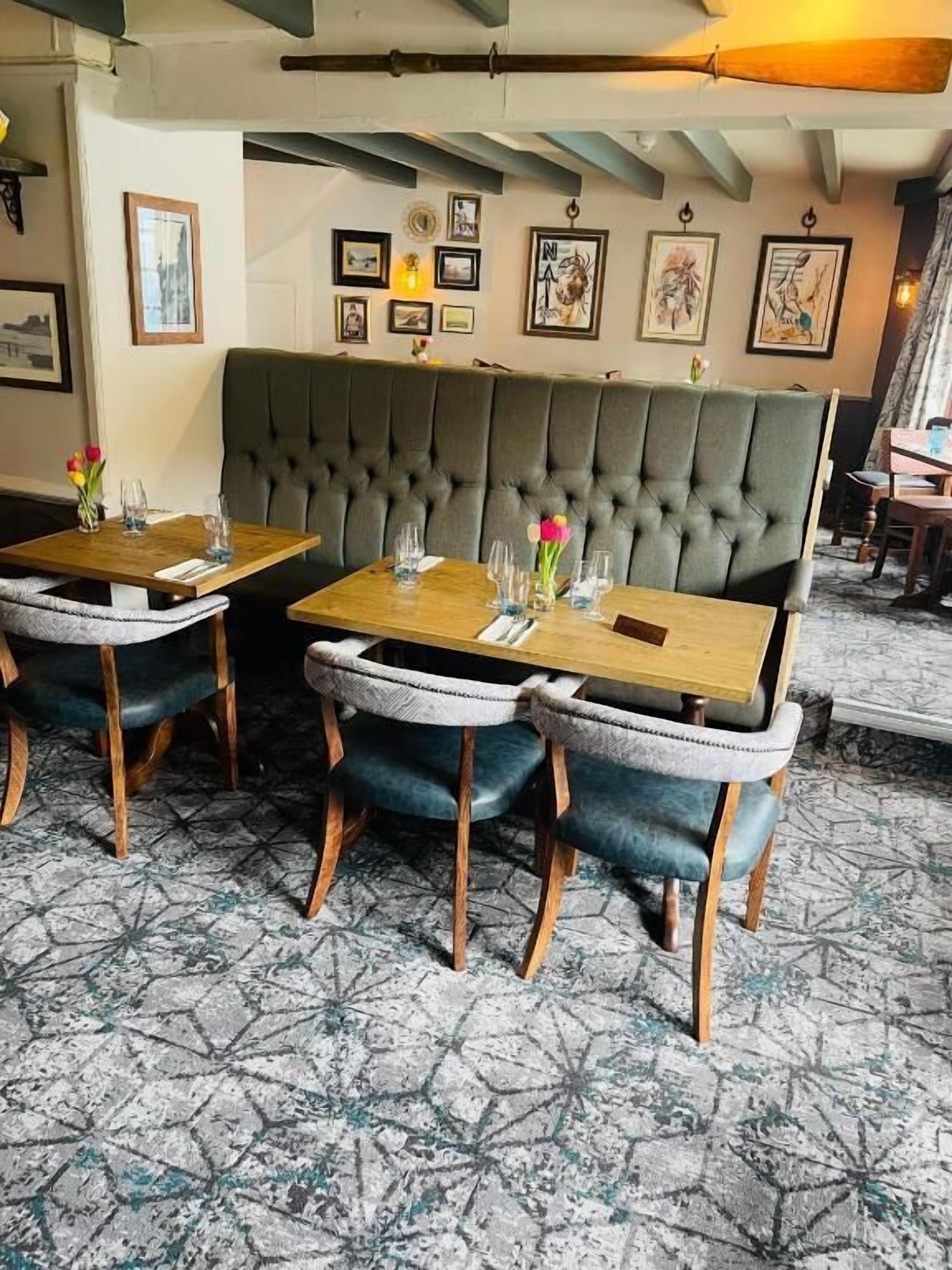 Un interior de restaurante con una zona central de mesas que incluye un largo banco con respaldo acolchado, mesas de madera y sillas tapizadas.