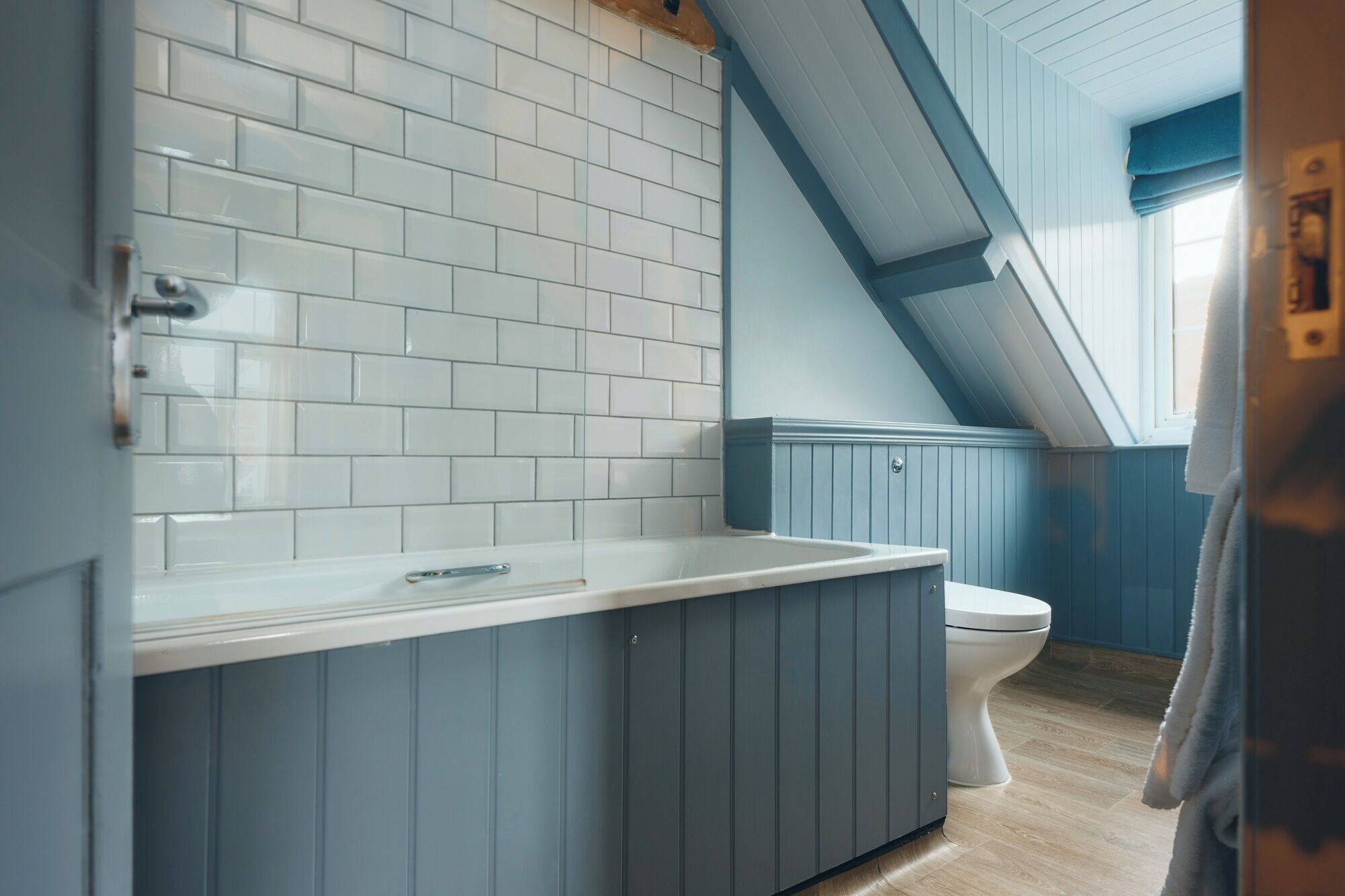 A bathroom with white tiled walls, a blue vanity, a white toilet, and a wooden floor.