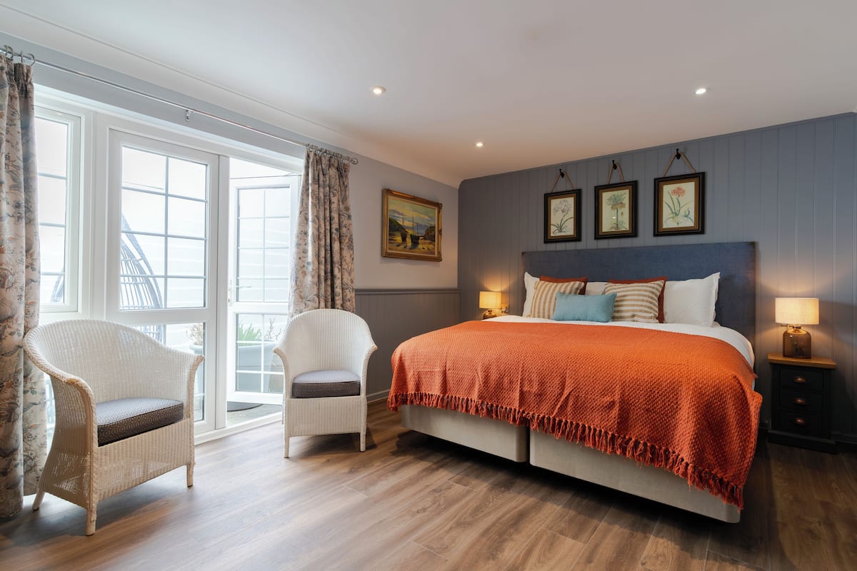 A bedroom with a large bed, a white chair, a window with curtains, and a painting on the wall.