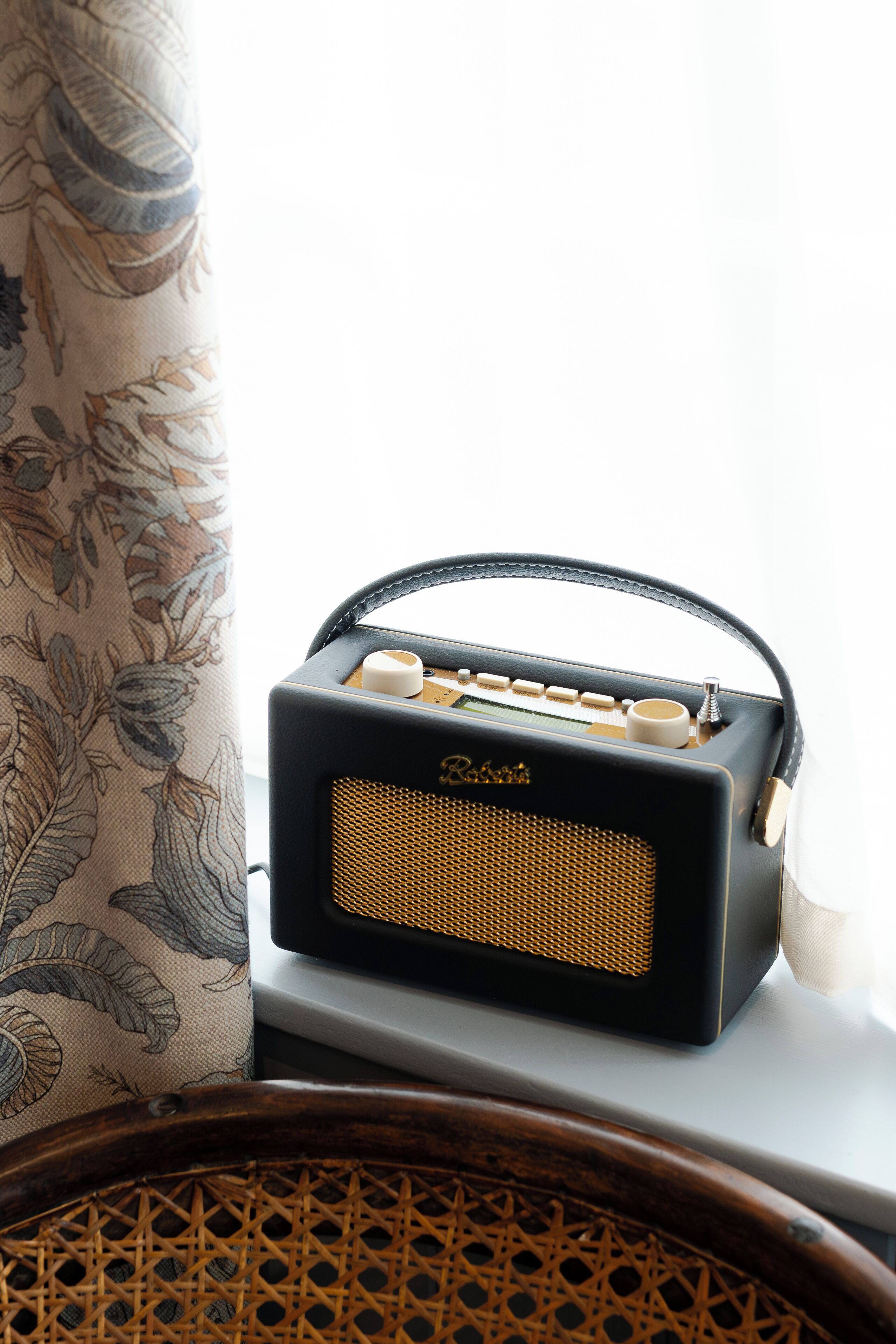 Une vieille radio posée sur une table, près d’une fenêtre ornée de rideaux à motifs floraux.