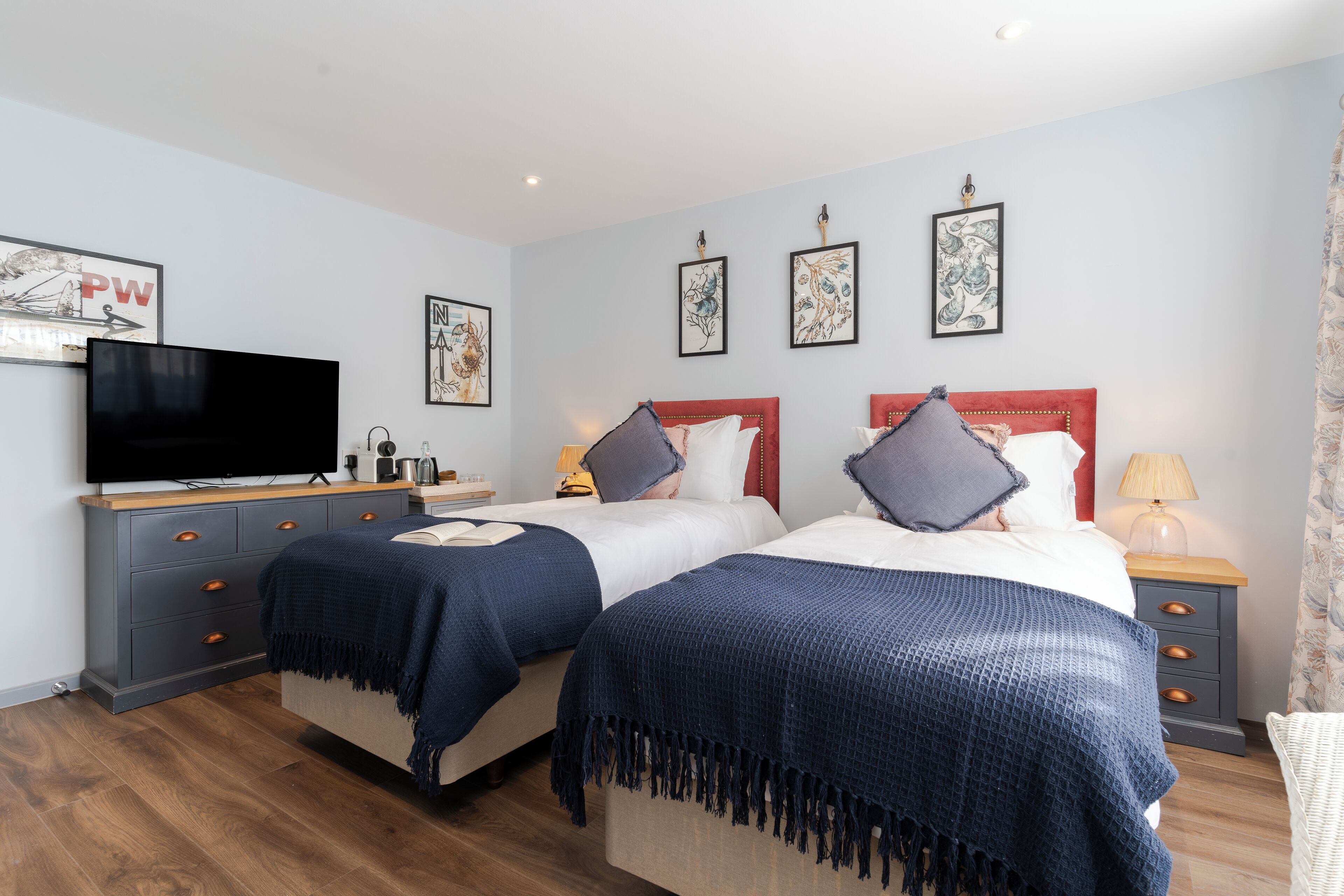 A hotel room with two beds, a TV, a bedside table with a lamp, and artwork on the walls.