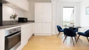 Una cocina moderna con armarios blancos, horno empotrado y microondas. Hay un comedor con mesa y sillas azules.