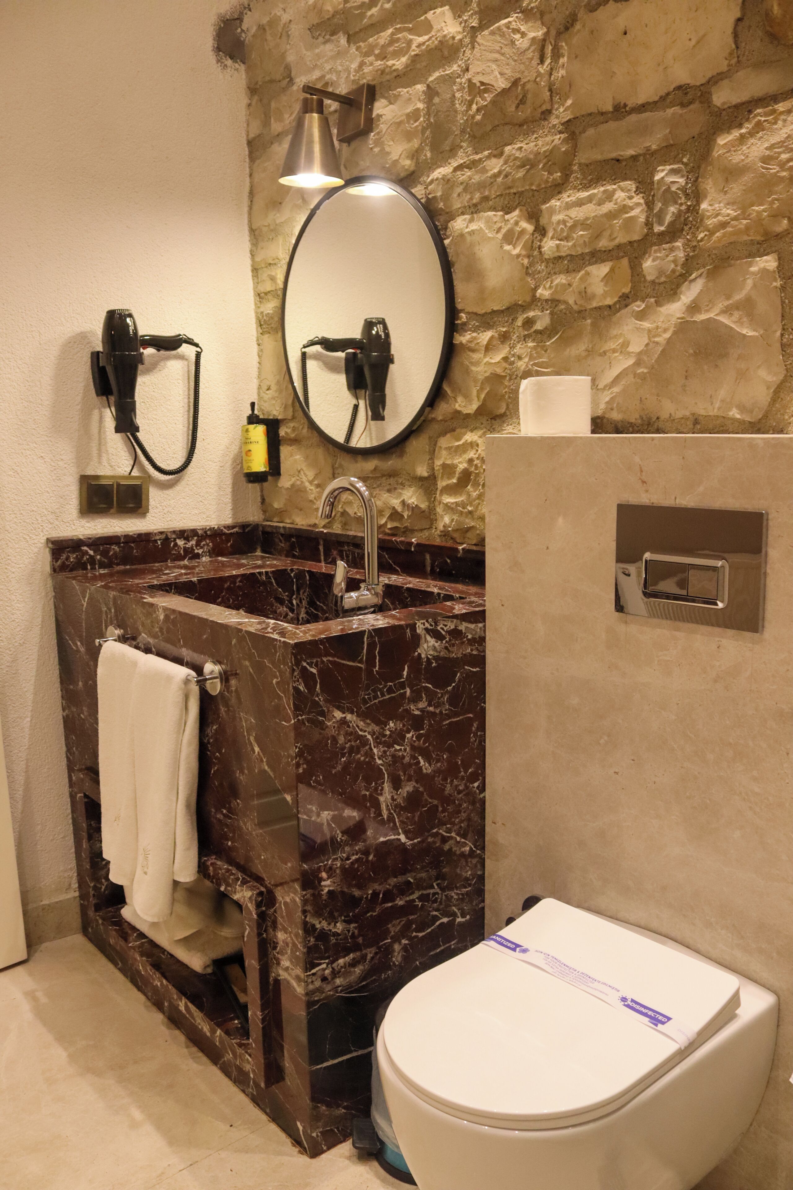 Un baño con lavamanos de mármol, inodoro, espejo y lámpara de pared.