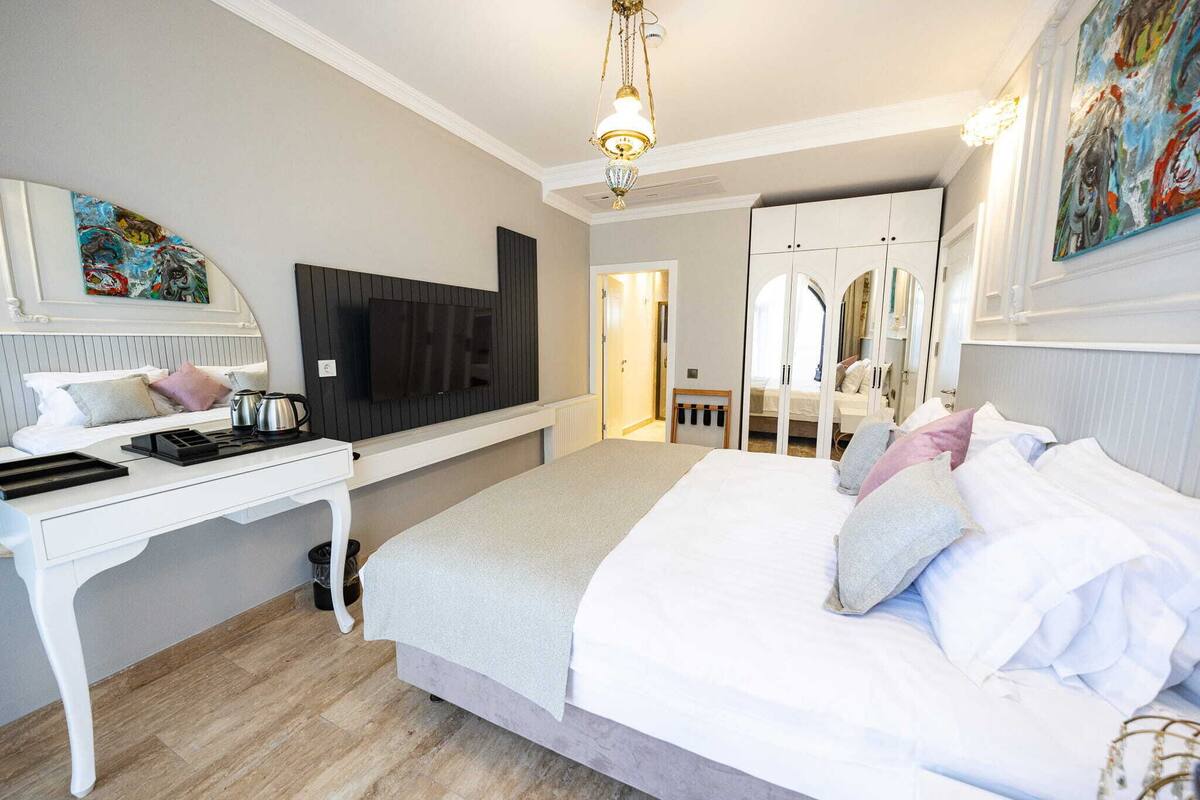 A bedroom with a bed, a desk, a television, a mirror, and a door leading to another room.