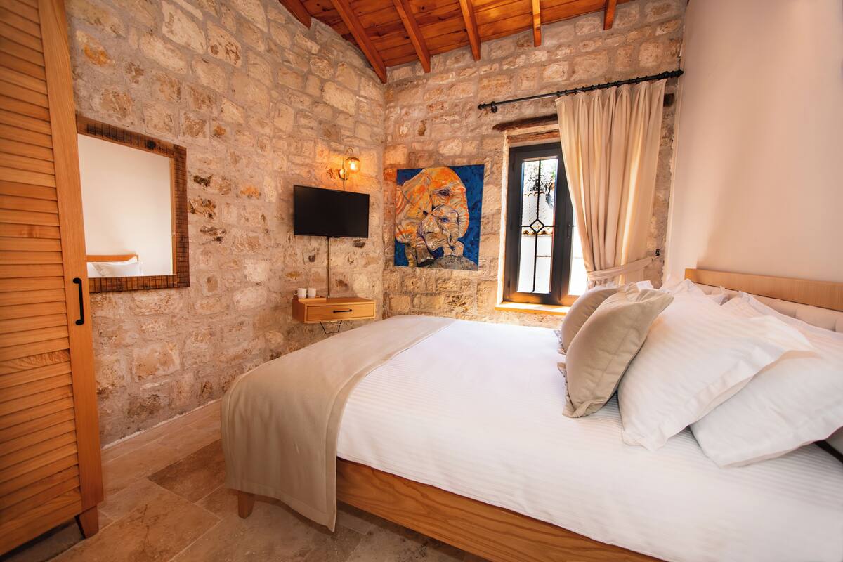 A bedroom with stone walls, a wooden bed frame, a mounted TV, and a window with curtains.