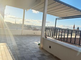 Área externa coberta com piso de azulejos, corrimão e vista para um edifício distante com um minarete.