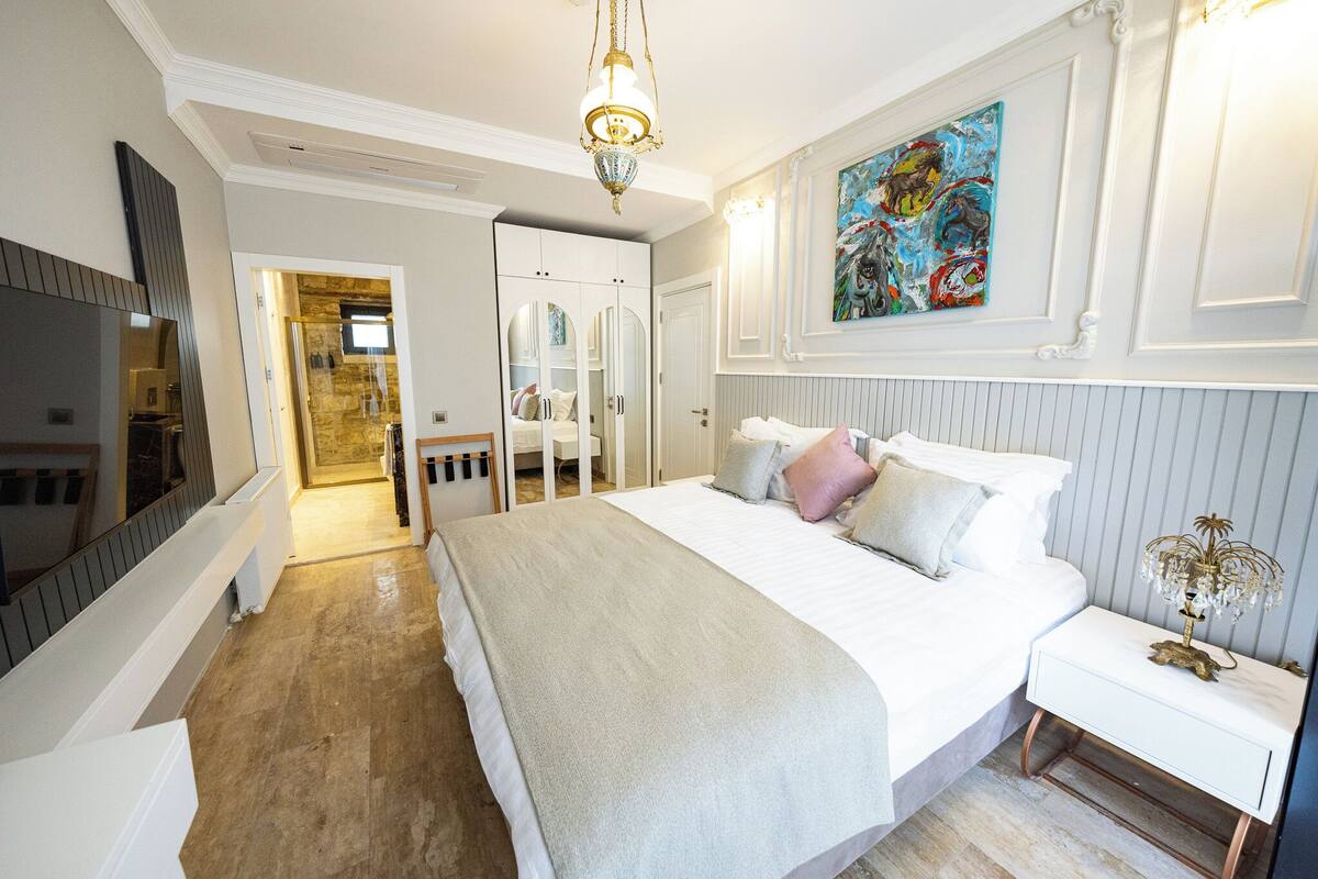A bedroom with a large bed, a chandelier, a painting, and a wardrobe.
