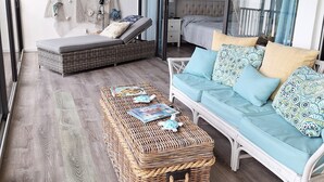 Un balcon spacieux avec une petite table basse en osier, un canapé blanc agrémenté de coussins à motifs, un ventilateur de plafond et une vue sur la mer à travers de grandes fenêtres.