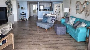 Un salon comprenant un téléviseur, un canapé, un pouf et un fauteuil en osier. Au fond, on aperçoit une cuisine avec des placards et un coin repas.
