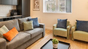 A modern living room with a grey sofa, two armchairs, and a coffee table. There is a kitchen area with black cabinets and a microwave. A window provides natural light and a view of trees. - Host Stay Princes Place Hideaway IV (Liverpool)