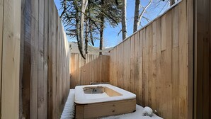 Zona d'spa de fusta amb banyera d'hidromassatge, envoltada de terra i arbres coberts de neu.