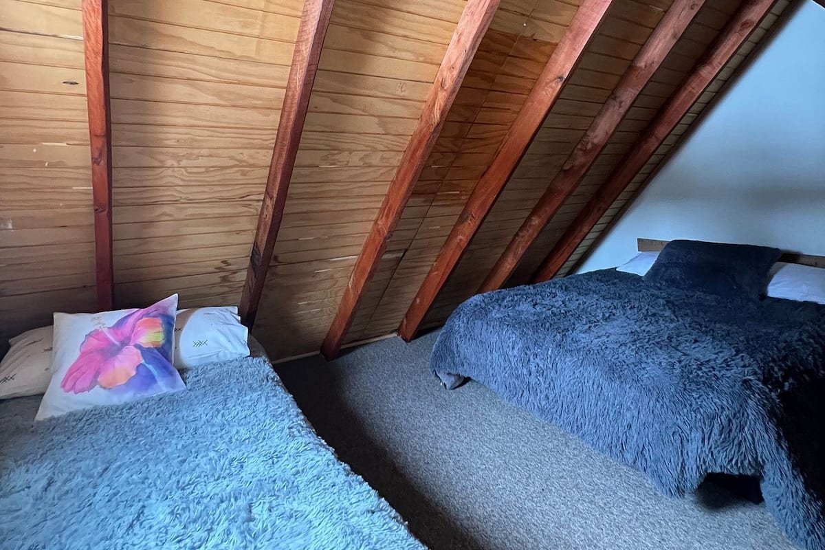 Een slaapkamer in een loft met zichtbare houten balken, twee bedden met zachte dekbedden, en een witte plafondlamp.
