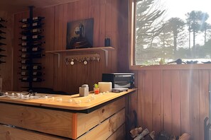 Een houten cabin interieur met een keukenblad, wijnflessen, een magnetron en uitzicht op bomen door een raam.