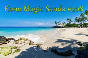 Una playa con agua turquesa cristalina, arena blanca y rocas. Se ven palmeras y un edificio al fondo.