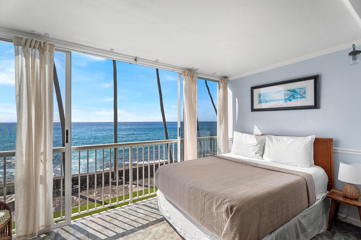Een slaapkamer met een groot raam met uitzicht op de oceaan, een bed met witte lakens en een lijst met een afbeelding aan de muur.