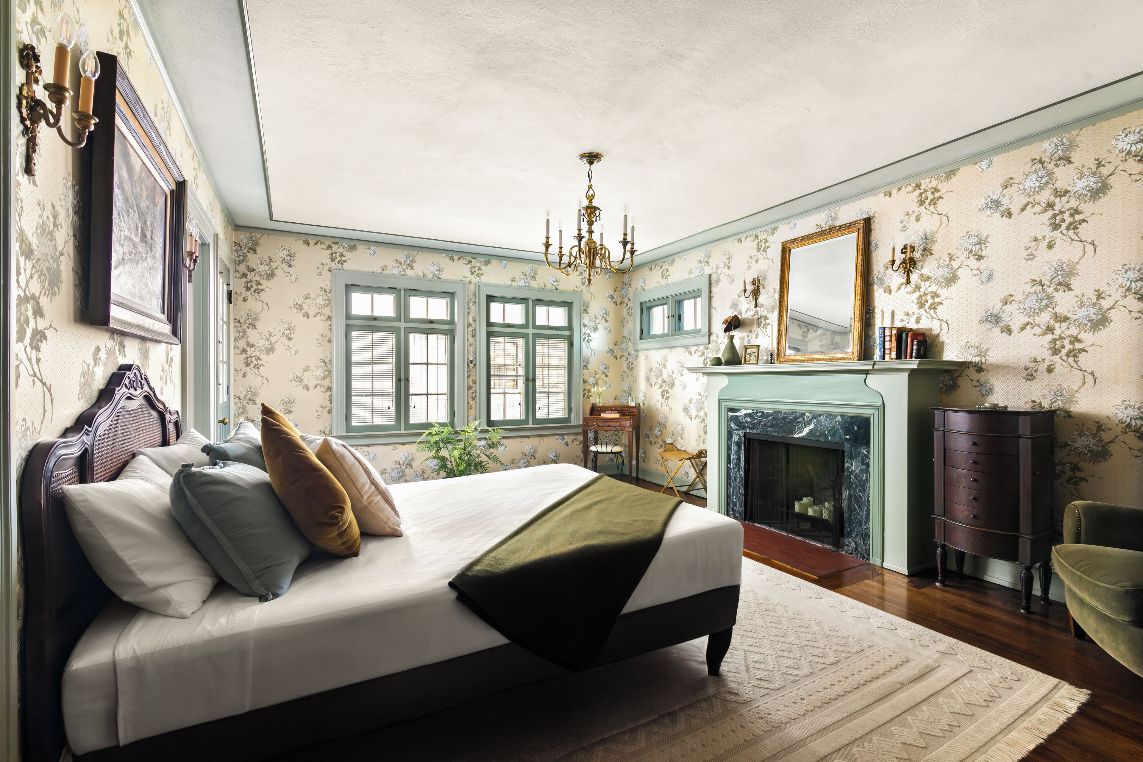 A bedroom with a large bed, a fireplace, a mirror, and floral wallpaper.