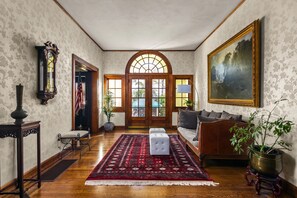 Un salon spacieux doté d’un parquet, d’un tapis à motifs rouges, d’un canapé, d’un tableau et d’une grande fenêtre donnant sur un paysage verdoyant.