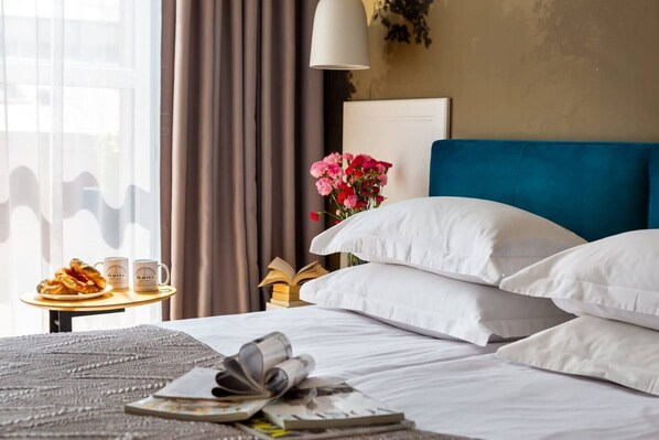 A hotel room with a bed, pillows, a bedside table with books and a plate of pastries, a lamp, and a window with curtains.