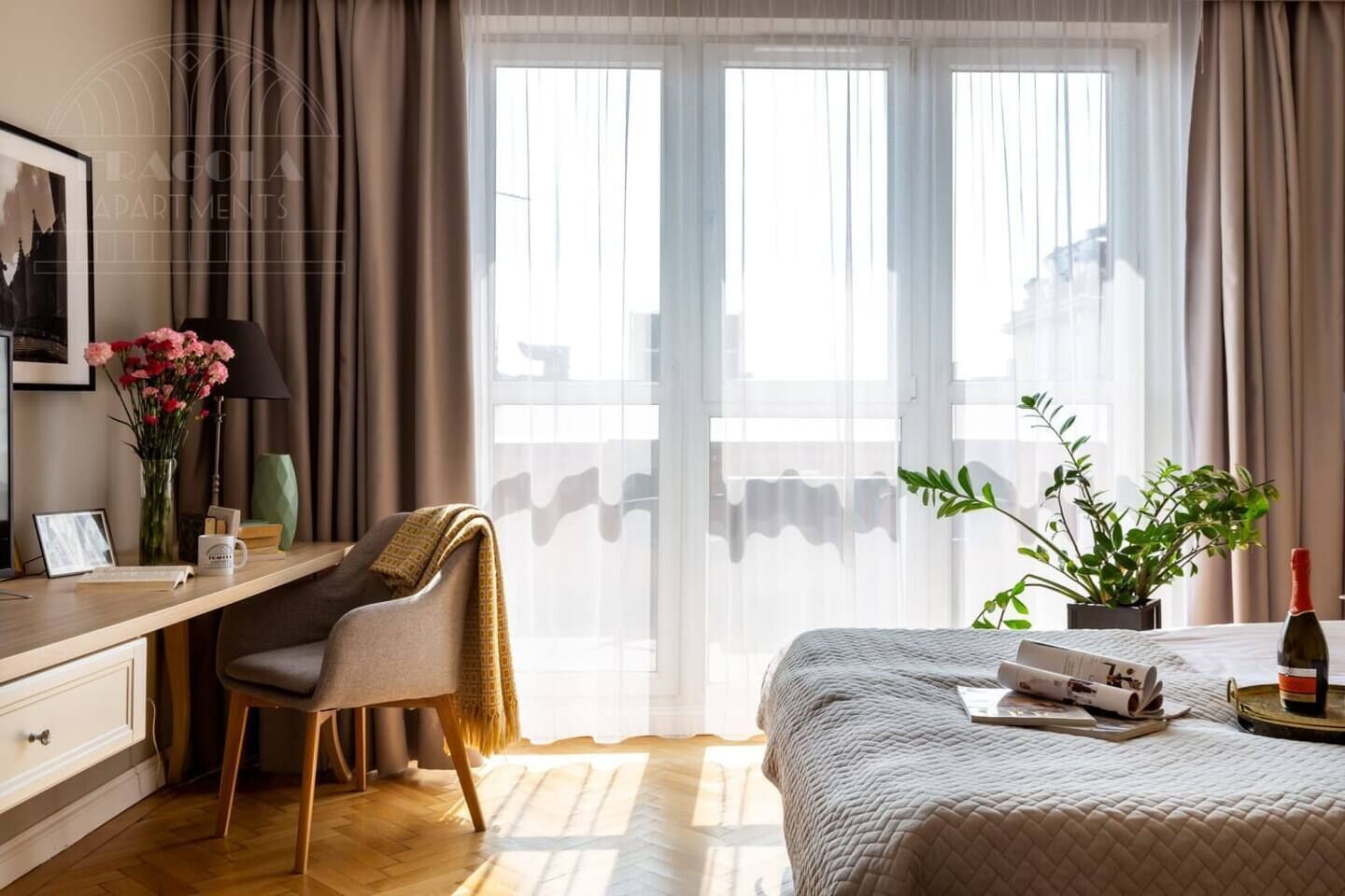A hotel room with a bed, a desk with a chair, a window with curtains, and a potted plant.