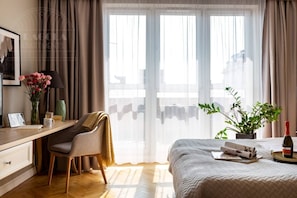 A hotel room with a bed, a desk with a chair, a window with curtains, and a potted plant.