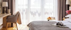 Habitación de hotel con cama, escritorio, silla, mesita con un plato de comida y una taza, y cortinas translúcidas.