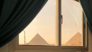 A room with a bed, a window with curtains, and a view of pyramids. - Pyramids Third Eye (Giza)