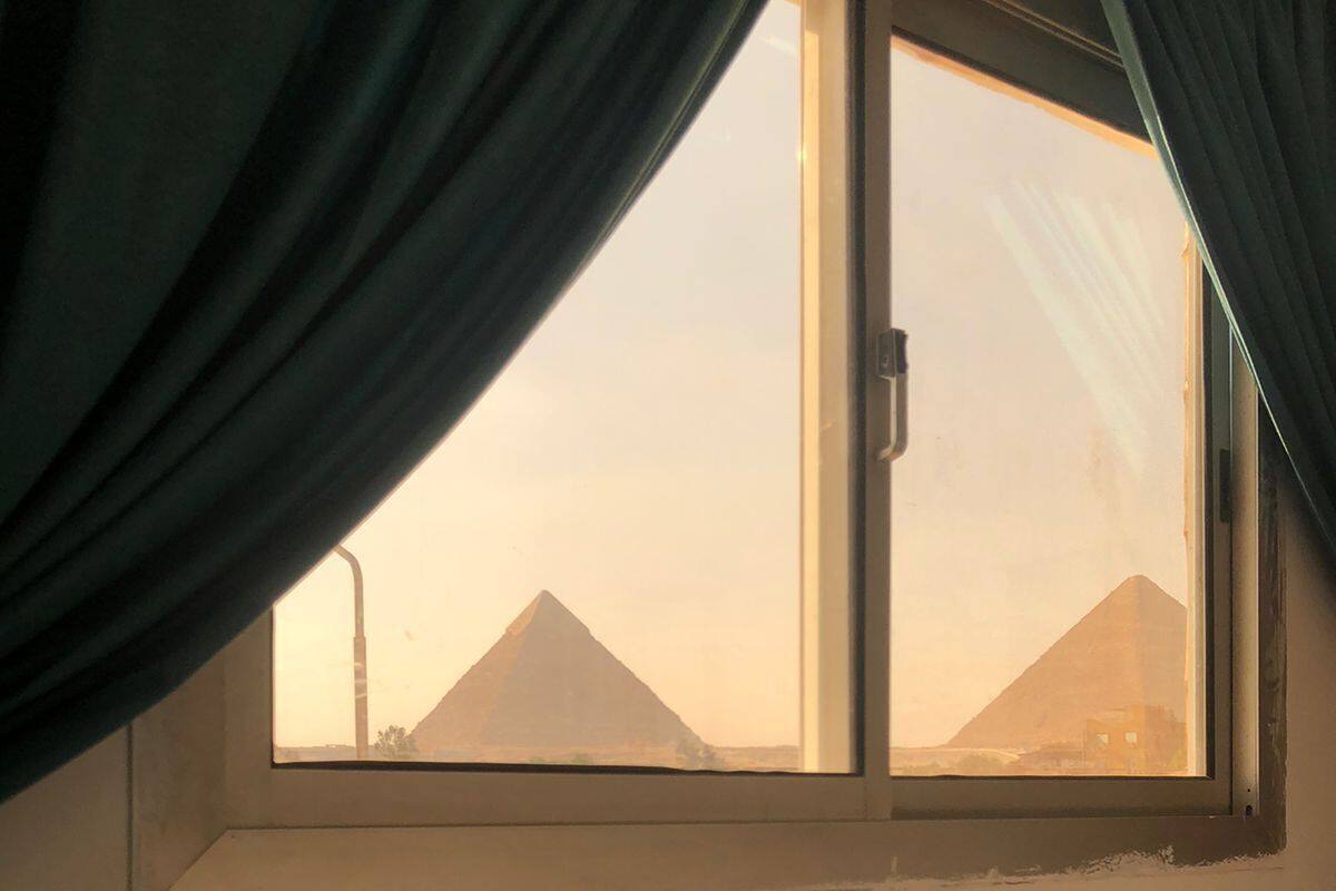 Ein Zimmer mit einem Bett, einem Fenster mit Vorhängen und Blick auf Pyramiden.