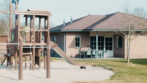 Un terrain de jeux avec un toboggan et des structures d’escalade, une maison en briques avec un toit en tuiles et un petit patio avec du mobilier d’extérieur.