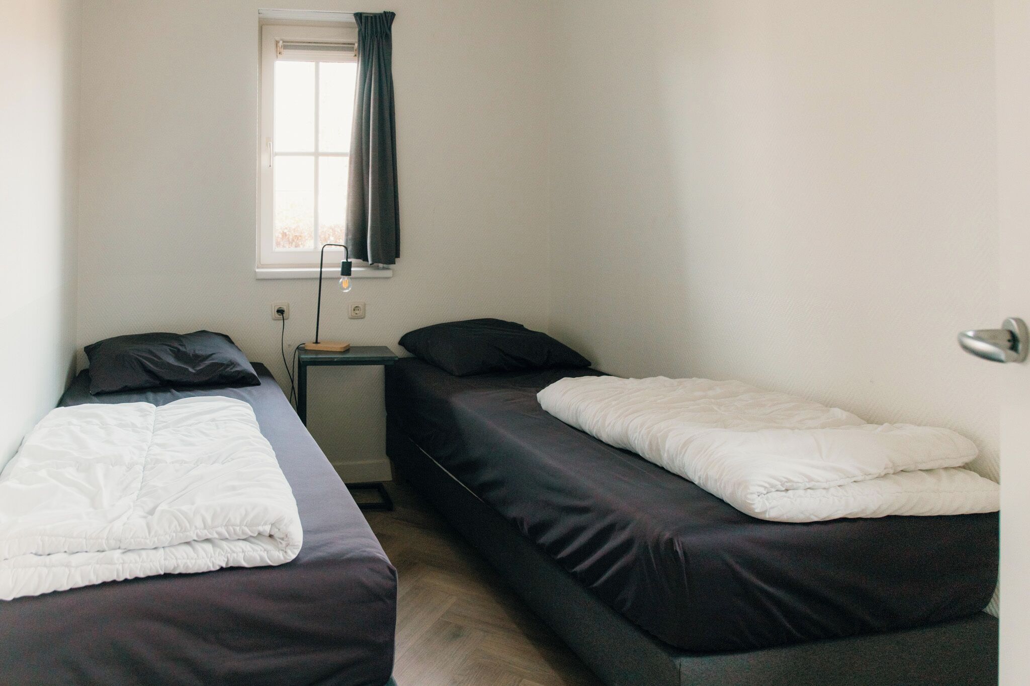 Een kleine, eenvoudige kamer met twee eenpersoonsbedden, een raam met gordijn en een nachtkastje met lamp.