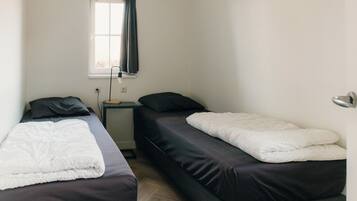 Een kleine, eenvoudige kamer met twee eenpersoonsbedden, een raam met gordijn en een nachtkastje met lamp.