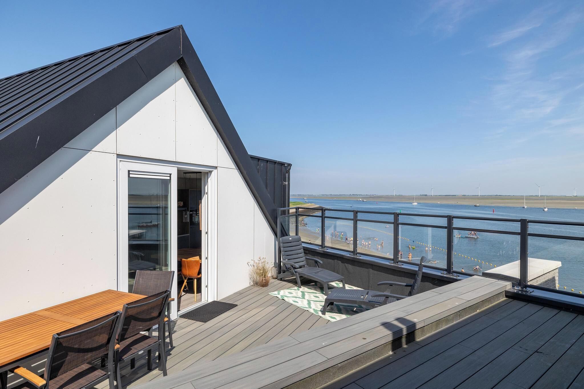 Een modern terras met een houten tafel en stoelen, een glazen deur die leidt naar een binnenruimte, en uitzicht op zee met schepen in de verte.