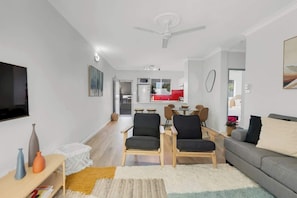 A modern living room with a flat-screen TV, two armchairs, a sofa, a dining area with a table and chairs, and a kitchen in the background.